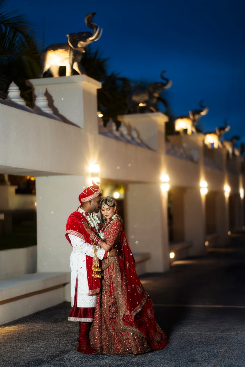 Newlyweds in red ceremonial dress beneath illuminated elephant sculptures